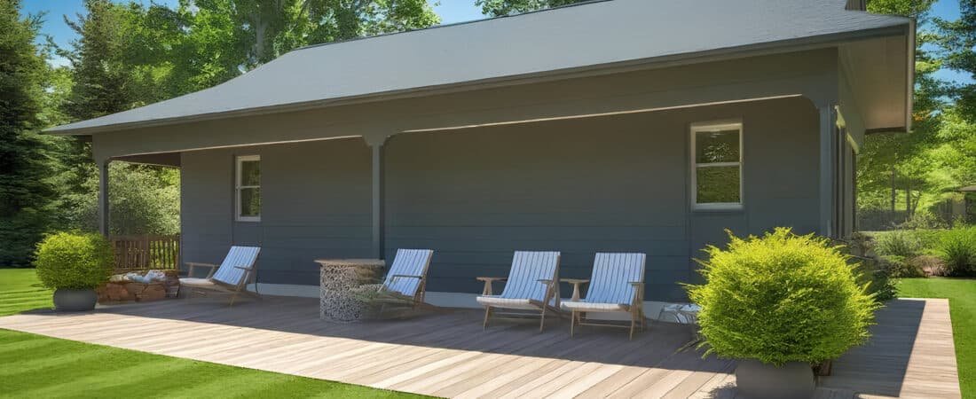 A modern farmhouse with dark metal siding and a large wooden porch featuring white rocking chairs, set on a lush green lawn under a clear blue sky