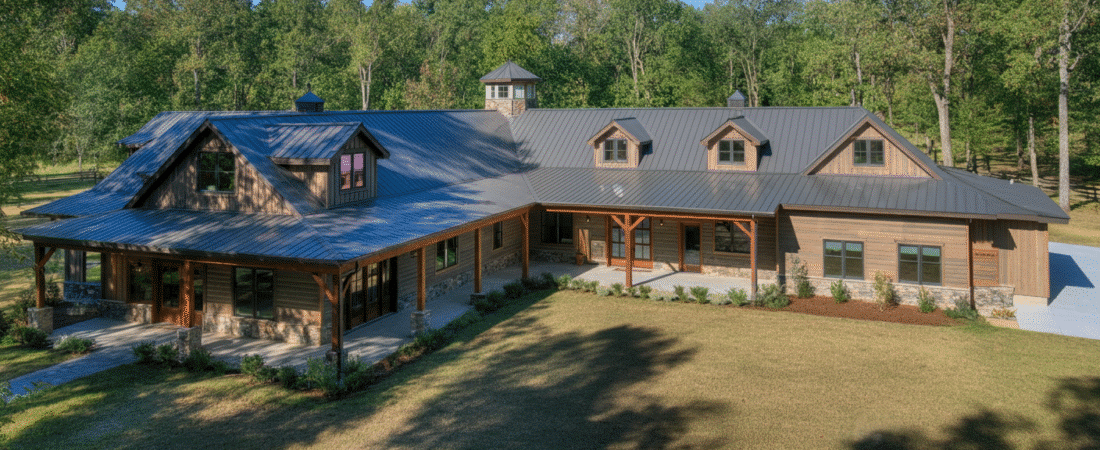 Photograph of a large, modern rustic house nestled against a lush forest, showcasing its expansive design, natural materials, and serene outdoor setting under a clear blue sky.