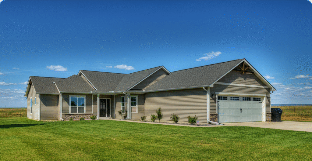 Photograph of a modern, well-maintained ranch-style house with tan siding and a gray roof, set against a vibrant green lawn and clear blue sky in a serene rural landscape.
