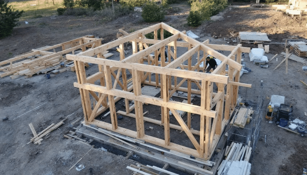 image - Ranch Style Homes USA The image depicts a construction site showcasing a farmhouse frame with exposed timber elements, highlighting the rustic charm and modern features characteristic of contemporary farmhouse architecture. The structure features gabled roofs and large windows, emphasizing the blend of traditional and modern aesthetics in its design.