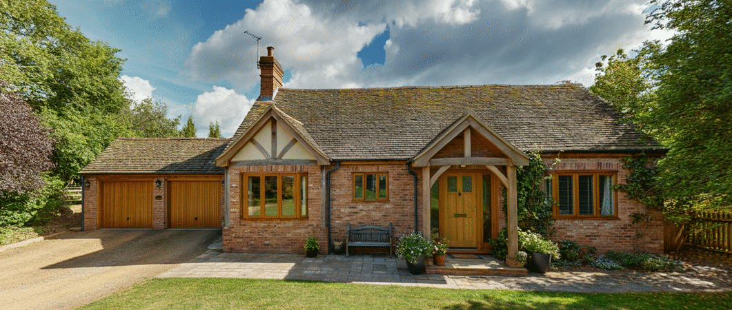 Photograph of a charming, traditional brick cottage with a double garage, lush green lawn, and mature trees under a partly cloudy sky, evoking warmth and tranquility.